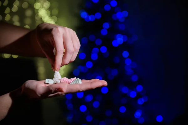 Close-up de mãos femininas. Uma mulher segura um punhado de marshmallows na forma de uma árvore de Natal no fundo das luzes de Natal . — Fotografia de Stock
