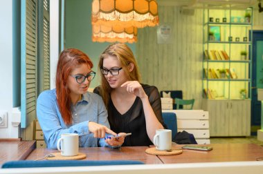 İki heyecanlı genç kız cep telefonu kullanıyor, bir kafede oturuyor ve parmakla işaret ediyor. Gözlüklü kızıl saçlı kadın akıllı telefon kız arkadaşının komik fotoğraflarını gösteriyor. Kahve içerler..