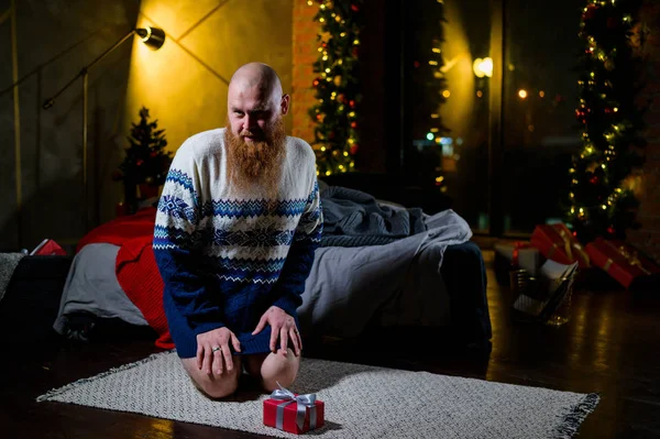 Un homme avec une longue barbe rouge est assis sur le sol sans pantalon dans un pull d'hiver. homme tient un cadeau enveloppé sur le fond des décorations et des lumières du Nouvel An. Arbre de Noël. Parodie, humour. — Photo