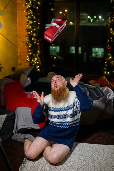 A man with a long red beard sits on the floor without pants in a winter sweater. man is holding a wrapped present on the background of New Year decorations and lights. Christmas tree. Parody, humor. — Stock Photo, Image
