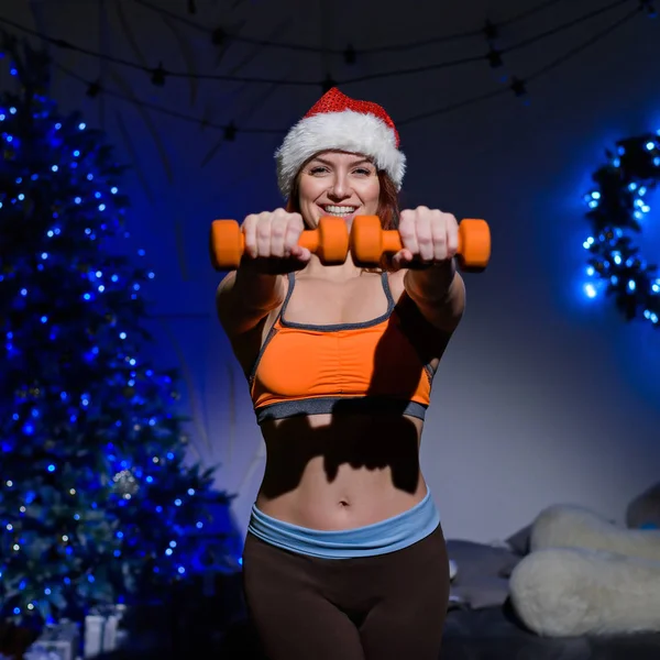 Atlet wanita berambut merah yang cantik dengan topi Santa Claus berpose di studio gelap. Wanita fit dalam pakaian olahraga memegang dumbel di tangannya dengan latar belakang pohon Tahun Baru . — Stok Foto