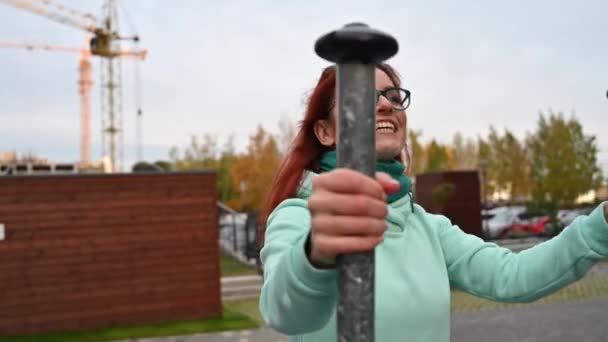 Una mujer pelirroja con gafas va a hacer deporte en un entrenador callejero. Una chica en una sudadera con capucha de menta se dedica a un simulador en el patio . — Vídeos de Stock