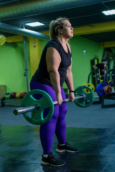 Fat European woman is engaged in fitness with a barbell in the gym. A fat girl does deadlift.