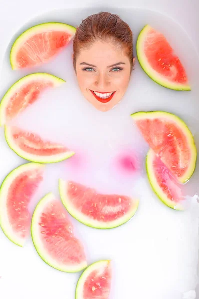 Portret van een mooie roodharige vrouw neemt een bad met melk en plakjes watermeloen. Spa behandeling voor huidverjonging en voeding. Rijpe plakjes van de grootste bes. — Stockfoto