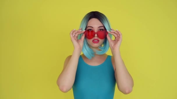 Beautiful girl in a blue wig and bikini posing on a yellow background. Woman with artificial hair and a swimsuit. heart-shaped sunglasses — Stok video