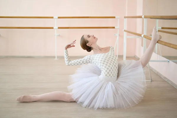 Krásná baletka v těle a bílá tutu cvičení v taneční třídě a posezení v rozštěpení. Mladý ohebný tanečník pózující ve špičatých botách. — Stock fotografie