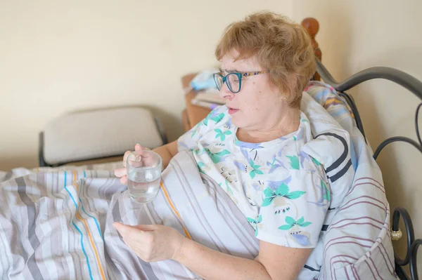 Sick old woman is lying at home in bed. A pensioner takes a cure for the disease and drinks a glass of water. — 스톡 사진