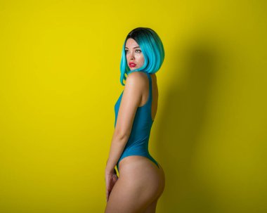 Mavi peruklu ve sarı arka planda poz veren parlak bikinili güzel bir kız. Yapay saçlı ve mayolu bir kadın kalçalarına vurgu yapıyor..