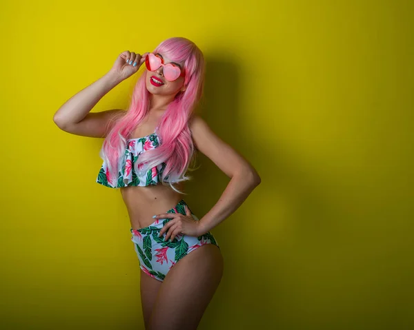 Menina bonita em uma peruca rosa e biquíni colorido posando em um fundo amarelo. Mulher com cabelos longos artificiais e óculos rosa na forma de corações . — Fotografia de Stock