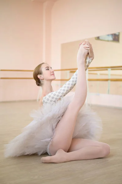 Bela bailarina em corpo e tutu branco está treinando em uma aula de dança. Jovem dançarina flexível posando em sapatos pontiagudos sentados no chão. — Fotografia de Stock