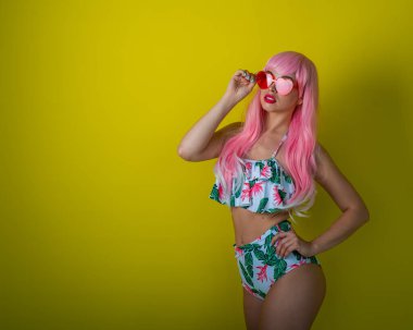 Pembe peruklu ve sarı arka planda poz veren renkli bikinili güzel kız. Yapay uzun saçlı ve kalp şeklinde pembe gözlüklü bir kadın..