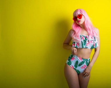 Pembe peruklu ve sarı arka planda poz veren renkli bikinili güzel kız. Yapay uzun saçlı ve kalp şeklinde pembe gözlüklü bir kadın..