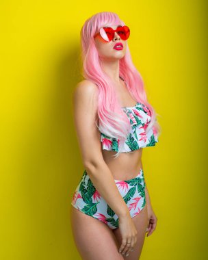 Pembe peruklu ve sarı arka planda poz veren renkli bikinili güzel kız. Yapay uzun saçlı ve kalp şeklinde pembe gözlüklü bir kadın..