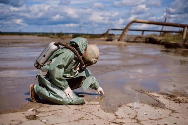 Bir bilim adamı kimyasal atık salınımından sonra nehirden su örneği alır. Solunum cihazına bağlı bir adam ve radyasyondan korunan yeşil bir giysi. Su analiz uzmanları. Ekolojik felaket.
