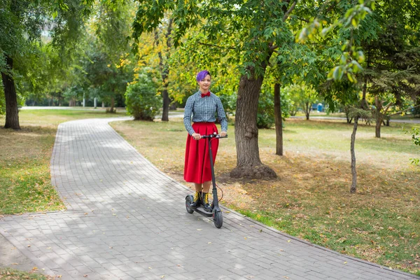 Mor saçlı genç bir kadın parkta elektrikli scooter kullanıyor. Ekose gömlek, uzun kırmızı etek ve papyon şık kız modern bir cihaz üzerinde şehir etrafında sürme. Hipster.