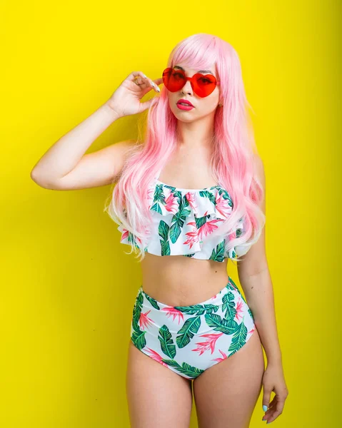 Pembe peruklu ve sarı arka planda poz veren renkli bikinili güzel kız. Yapay uzun saçlı ve kalp şeklinde pembe gözlüklü bir kadın..