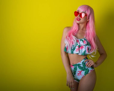 Pembe peruklu ve sarı arka planda poz veren renkli bikinili güzel kız. Yapay uzun saçlı ve kalp şeklinde pembe gözlüklü bir kadın..
