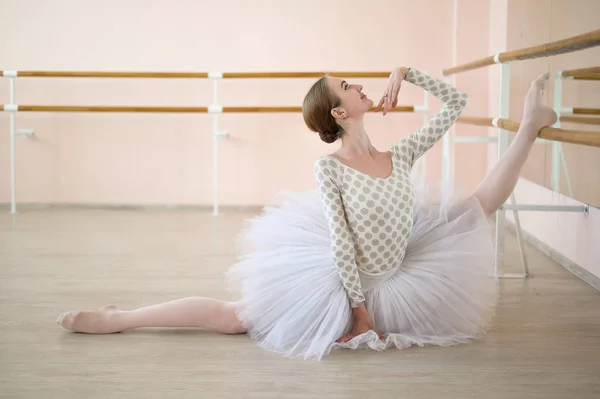 Bela bailarina em corpo e tutu branco está treinando em uma aula de dança. Jovem dançarina flexível posando em sapatos pontiagudos sentados no chão. — Fotografia de Stock