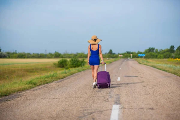 Een vrouw met een hoed en een grote tas loopt langs de weg. Meisje in een blauwe strakke jurk met een paarse koffer op de baan. Brunette met een sportfiguur op de weg. — Stockfoto