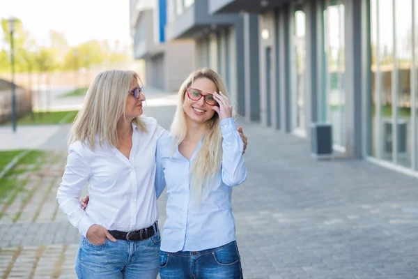 Een mooie blonde met bril en haar oudere moeder lopen samen. Moederdag. Mooie oude vrouw is een oudere burger en haar volwassen dochter. Gelukkige vrouwen van verschillende generaties. — Stockfoto