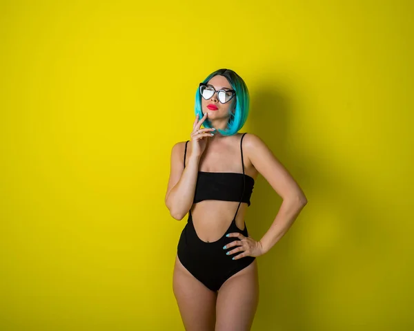 Beautiful woman in a black bikini in a colored wig and sunglasses on a yellow background. Girl posing in the studio. — 스톡 사진