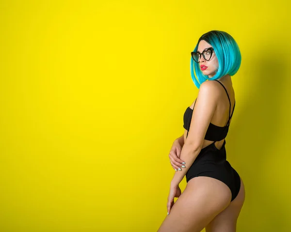 Mooie vrouw in een zwarte bikini met een gekleurde pruik en zonnebril op een gele achtergrond. Meisje poseren in de studio. — Stockfoto