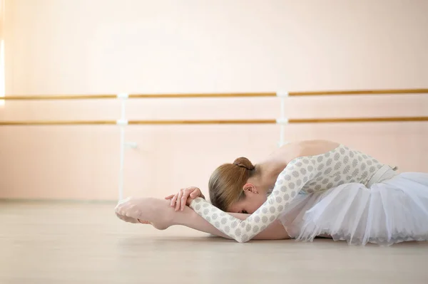 Bela bailarina em corpo e tutu branco está treinando em uma aula de dança. Jovem dançarina flexível posando em sapatos pontiagudos sentados no chão. — Fotografia de Stock