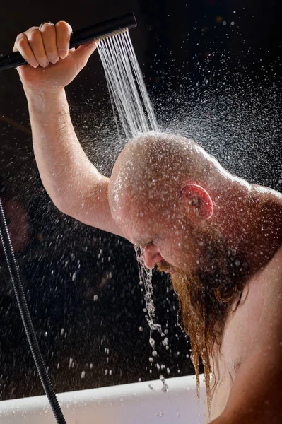 El macho toma una ducha. Un hombre brutal con barba roja está parado en el baño bajo el agua corriente y se lava. Spray scatter sobre un fondo negro. — Foto de Stock