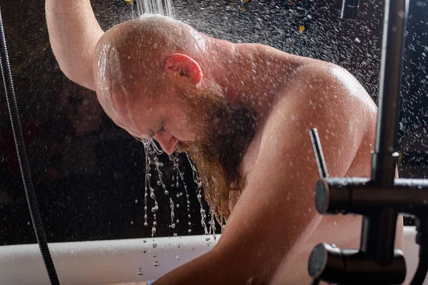Man tar en dusch. En brutal man med rött skägg står i badrummet under rinnande vatten och tvättar sig. Spraya scatter på en svart bakgrund. — Stockfoto