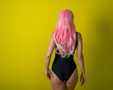 Uzun dalgalı yapay saçlı bir kadın siyah bir mayo giyiyor. Sarı arka planda kıvırcık pembe peruklu moda kızı..