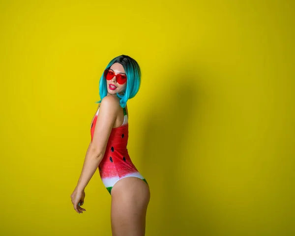 Portrait of a woman in a swimsuit with a picture of a watermelon and red sunglasses in the shape of hearts. Stylish girl in a colored short wig posing in the studio on a yellow background. — Stock Photo, Image