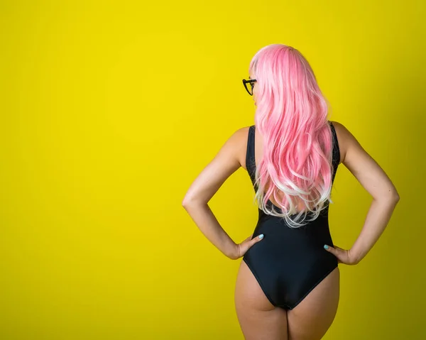 A woman with long wavy artificial hair is standing back in a black swimsuit. Fashion girl in a curly pink wig on a yellow background.
