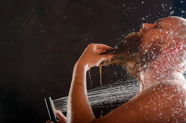 El calvo se baña. Un hombre brutal con barba roja está parado en el baño bajo un chorro de agua y se lava. Spray scatter sobre un fondo negro . — Foto de Stock
