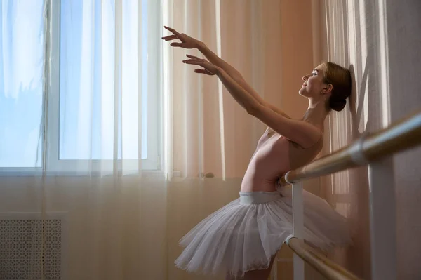 Piękna, pełna wdzięku balerina ćwiczy w holu na tle okna. Szczupła kobieta in tutu i szpiczaste buty w klasie tańca. — Zdjęcie stockowe