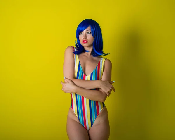 Beautiful woman in a striped swimsuit in a blue wig posing on a yellow background. Portrait of a glamorous girl. — Stock Photo, Image