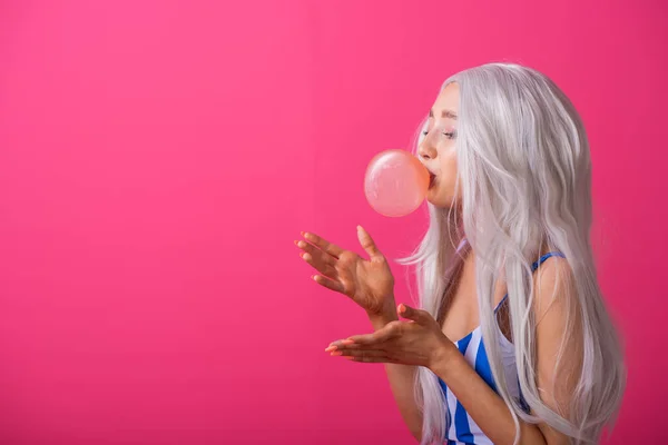 Portrait of a slender Asian woman in a blonde wig posing on a pink background. Cute girl in a striped swimsuit chewing gum and blowing bubbles. — 스톡 사진