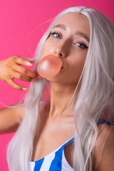 Portrait of a slender Asian woman in a blonde wig posing on a pink background. Cute girl in a striped swimsuit chewing gum and blowing bubbles. — 스톡 사진