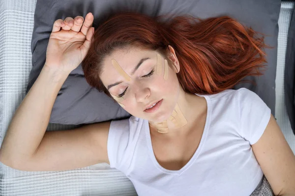 Fita Kineyzio no rosto de uma menina adormecida. Uma mulher está deitada em uma cama com manchas para rejuvenescimento facial . — Fotografia de Stock