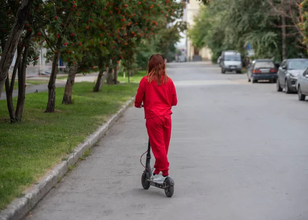 Κοκκινομάλλα κορίτσι σε μια κόκκινη φόρμα οδηγεί ένα ηλεκτρικό σκούτερ. Μια νεαρή γυναίκα με υπερμεγέθη ρούχα περιφέρεται γύρω από την πόλη και ακούει μουσική χρησιμοποιώντας ασύρματα ακουστικά. Θέα από πίσω. — Φωτογραφία Αρχείου