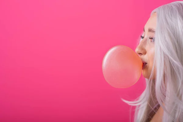 Портрет стройной азиатки в блондинистом парике, позирующей на розовом фоне. Милая девушка в полосатом купальнике жует жвачку и пускает пузыри. . — стоковое фото