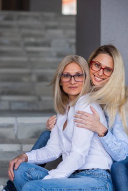 Gözlüklü güzel bir sarışın ve olgun annesi dışarıda merdivenlerde oturuyor. İyi giyimli yaşlı bir kadın yetişkin kızına sıkıca sarılır. Aşk ve aile değerleri. Anneler Günü.