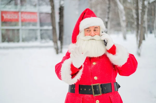 Здивований Санта Клаус використовує мобільний телефон на Північному полюсі. Санта Клаус стоїть в парку взимку і кличе смартфон. Сніг у грудні. — стокове фото