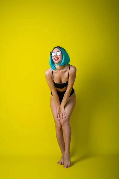 Beautiful woman in a black bikini in a colored wig and sunglasses on a yellow background. Girl posing in the studio. — 스톡 사진