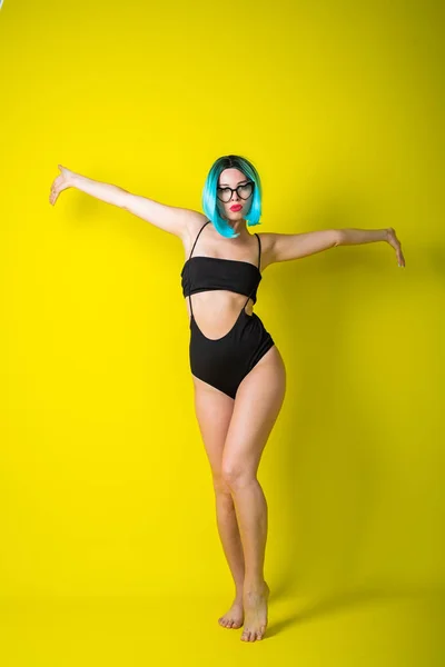 Belle femme en bikini noir avec une perruque colorée et des lunettes de soleil sur fond jaune. Fille posant dans le studio . — Photo