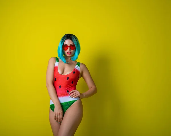 Portrait of a woman in a swimsuit with a picture of a watermelon and red sunglasses in the shape of hearts. Stylish girl in a colored short wig posing in the studio on a yellow background. — 스톡 사진