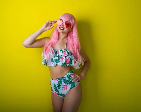 Schöne Mädchen in rosa Perücke und farbigem Bikini posiert auf gelbem Hintergrund. Frau mit künstlichen langen Haaren und rosa Brille in Herzform. — Stockfoto