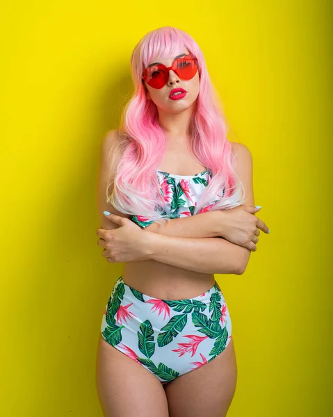 Pembe peruklu ve sarı arka planda poz veren renkli bikinili güzel kız. Yapay uzun saçlı ve kalp şeklinde pembe gözlüklü bir kadın..