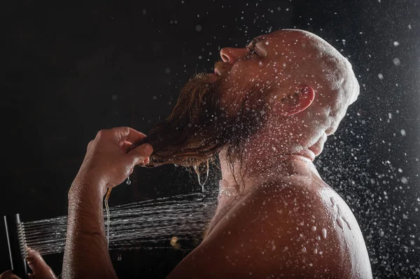 The bald guy takes a shower. A brutal man with a red beard is standing in the bathroom under a stream of water and washes. Spray scatter on a black background. — Stockfoto