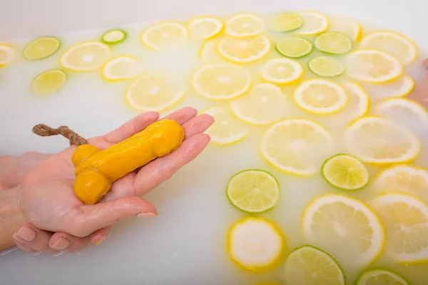 Fille prend un bain de lait avec des citrons et des citrons verts. Spa aux agrumes. Soins du corps. Blanchiment de la peau. Une femme se lave les mains avec du savon sous la forme d'un pénis masculin. Humour . — Photo