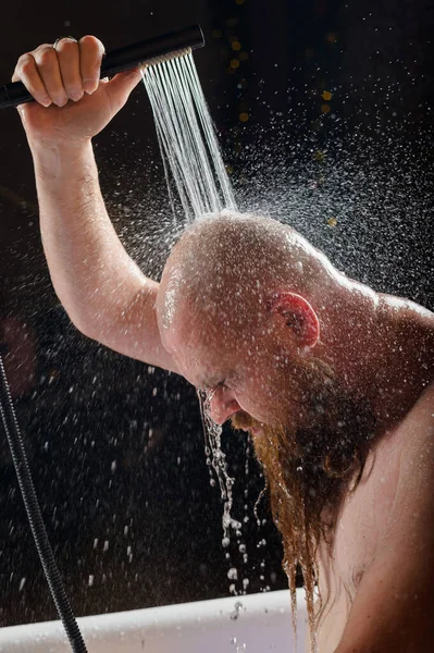 El macho toma una ducha. Un hombre brutal con barba roja está parado en el baño bajo el agua corriente y se lava. Spray scatter sobre un fondo negro. — Foto de Stock